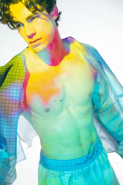 Young adult male in casual sporty fashion poses against a white backdrop with dynamic colorful lighting, showing his muscular upper body.
