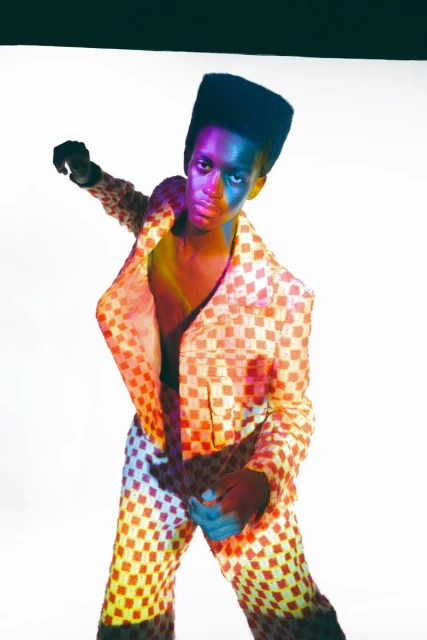 A stylish Black adult model in a multicolored patterned suit poses in a studio with creative lighting casting vibrant hues across the scene.