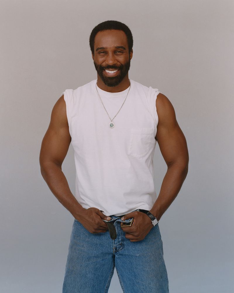 Portrait of a smiling Black man in a white tank top and blue jeans, standing against a neutral grey background in a photo studio.