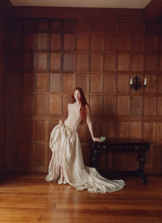 Elegant woman in a light-colored gown poses next to a dark wood table in a room with wood-paneled walls and hardwood floors.
