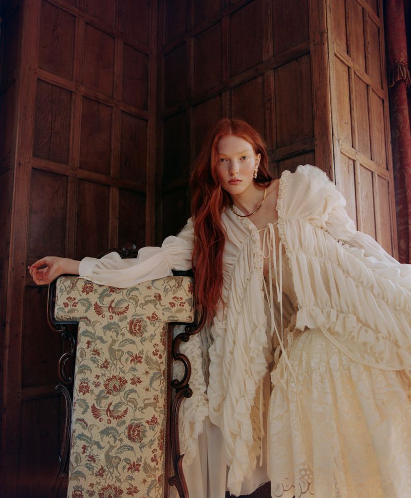 Red-haired woman in ruffled white dress poses with antique floral chair and wood paneling in editorial-style fashion photography indoors.
