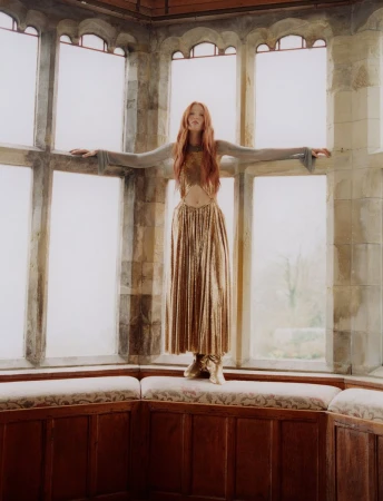 Full-length portrait of a woman in gold dress standing in a window alcove, arms outstretched, inside a castle-like room with stone and wood.