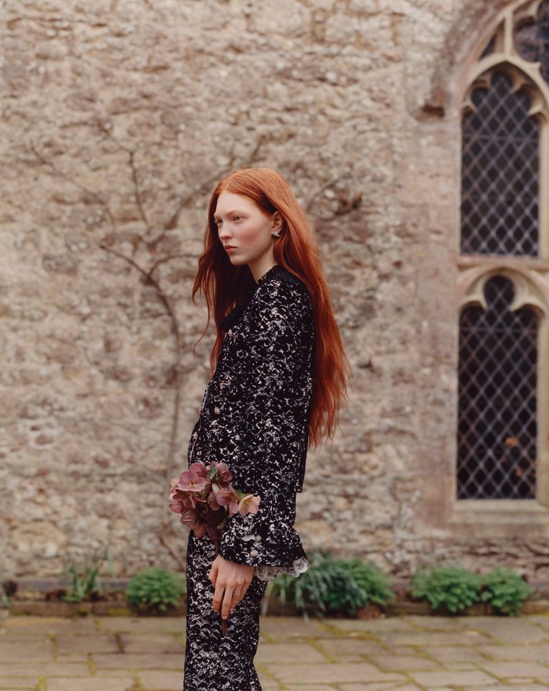 Red-haired woman in a black lace suit stands holding flowers in front of a stone building with arched windows on a cloudy day.