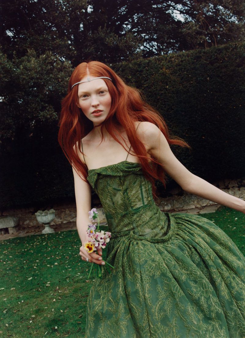 Red-haired woman in a green gown stands in a garden holding flowers, with trees and grass visible. Fashion, nature, and beauty are highlighted.