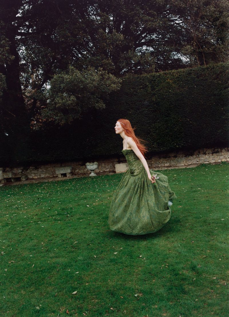 Red-haired woman in a formal green gown walking through a verdant garden with stone walls, mature trees, and lush green grass.