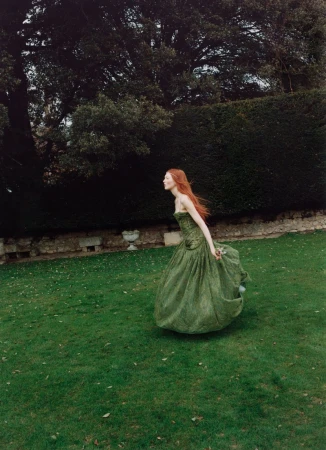 Red-haired woman in a formal green gown walking through a verdant garden with stone walls, mature trees, and lush green grass.