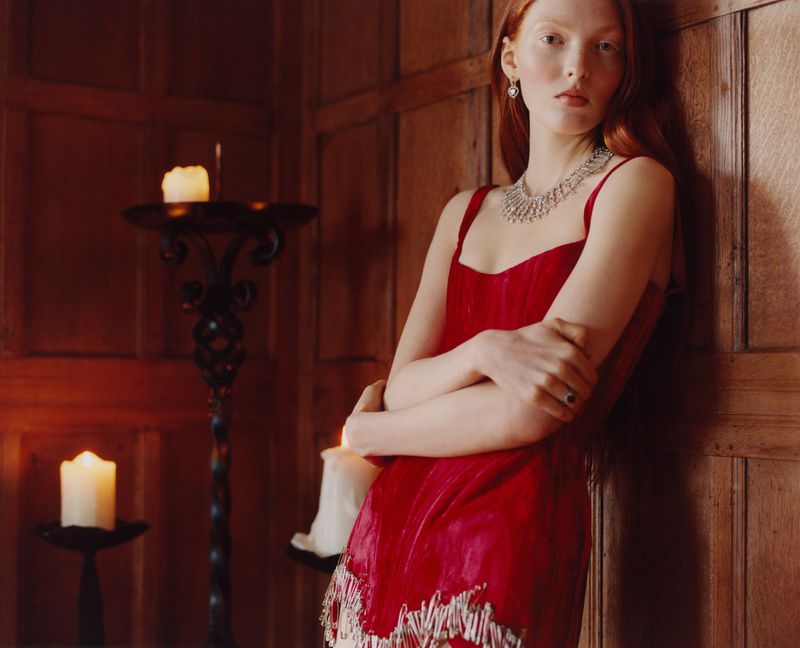 Elegant red-haired woman in a red velvet dress and jewelry leans against wood-paneled wall in a dimly lit, candlelit interior.