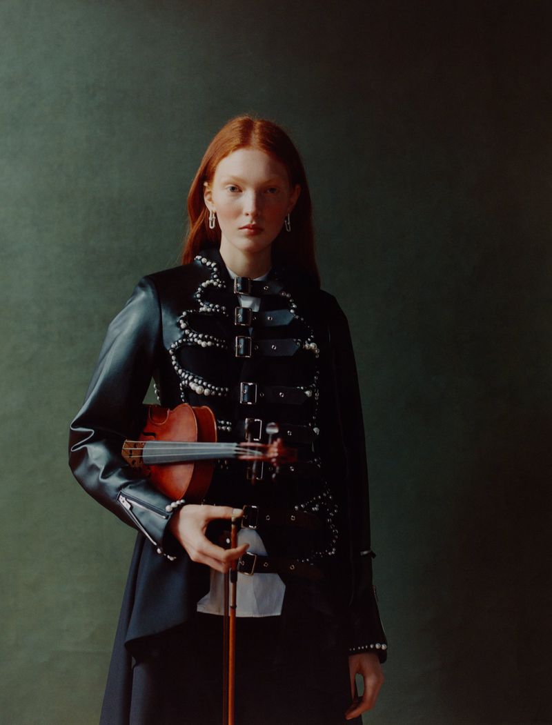 Red-haired woman in an embellished leather jacket poses indoors holding a violin and bow against a dark green backdrop.