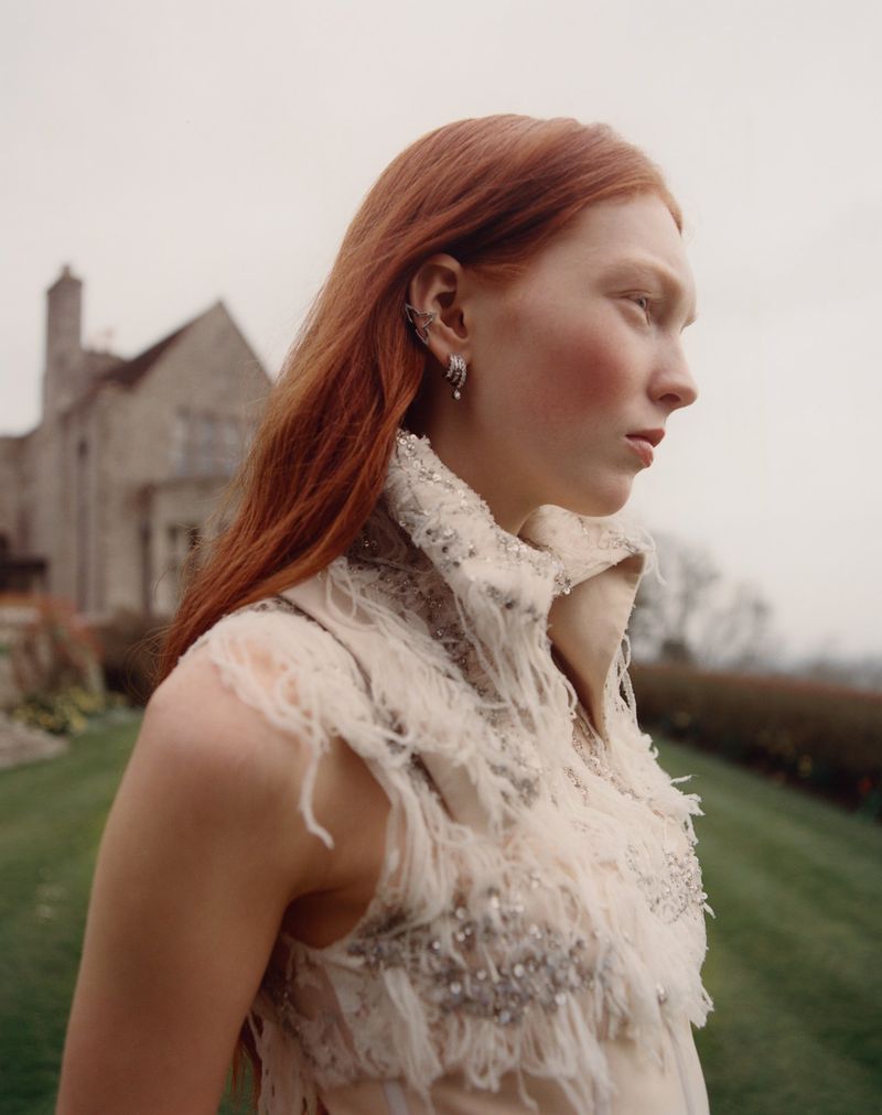 Profile of a red-haired woman in a textured white dress outdoors. She is wearing ear jewelry, with a large building and green grass in the background.