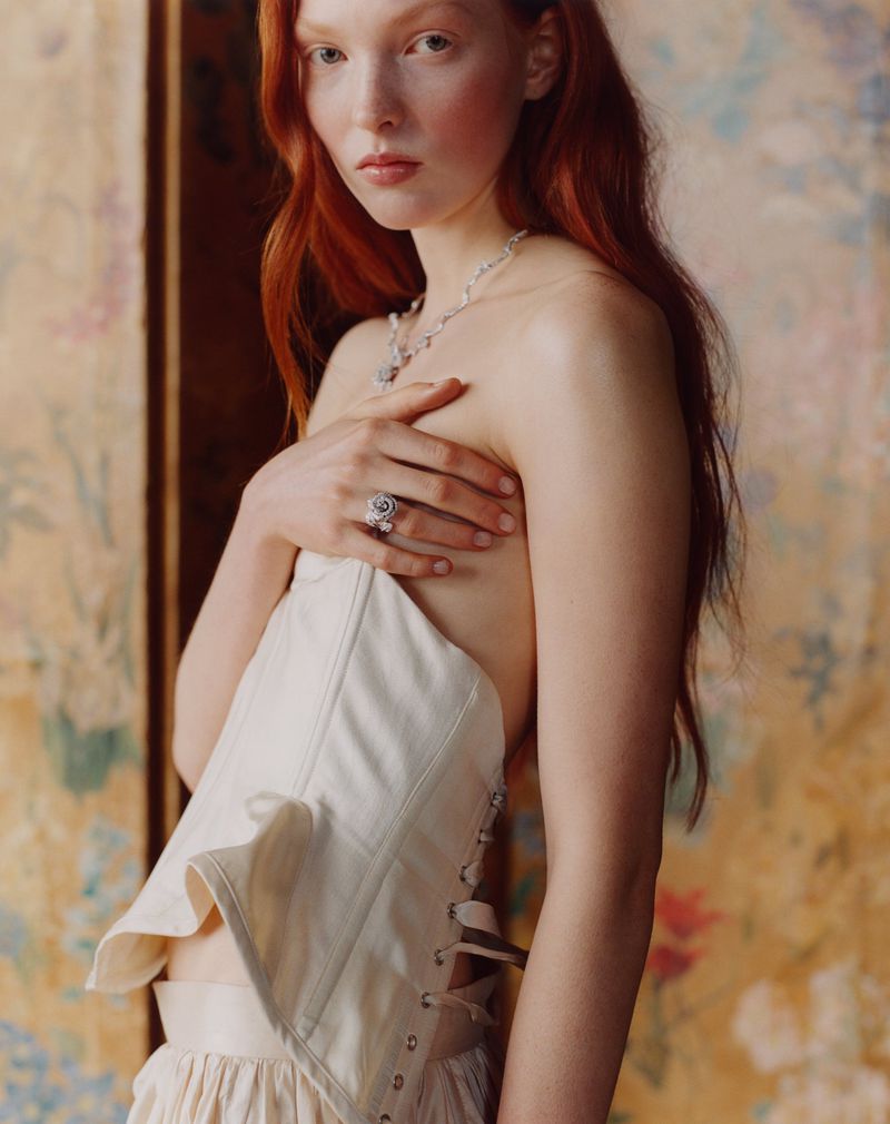 A red-haired woman poses indoors wearing a white corset top and skirt. She wears jewelry and is facing the camera.