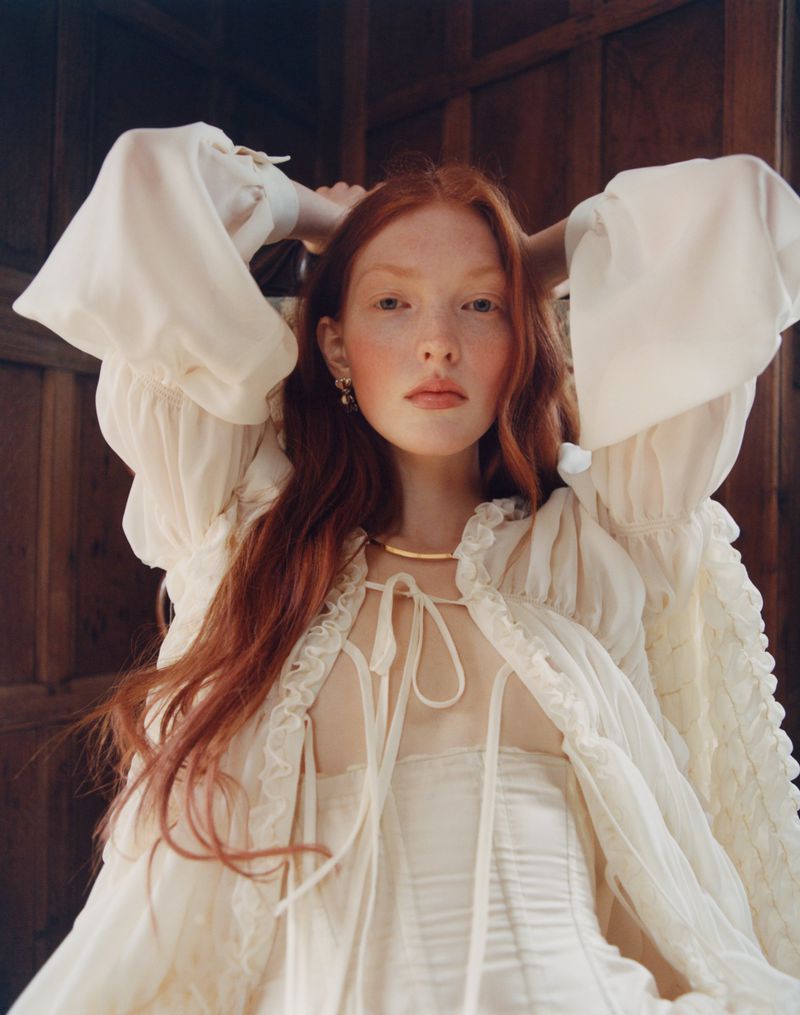 Portrait of a red-haired woman in a cream-colored sheer dress with a gold necklace posing in front of a wooden paneled wall.