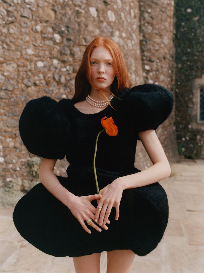 Red-haired woman in a sculptural black dress poses outdoors with an orange flower against a stone wall backdrop in a fashion editorial.