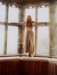 Model with long red hair wearing a gold pleated dress standing inside an ornate stone bay window
