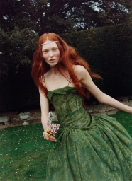 Model with long red hair wearing a green dress and holding small wildflowers in a garden setting