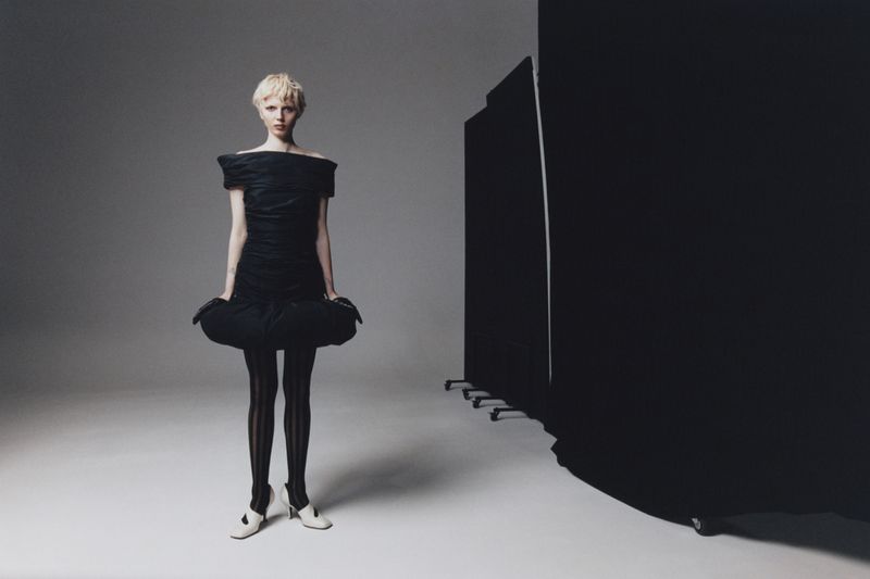 Fashion shot of a model in a black dress and striped tights, posing in a photo studio with a dark screen backdrop. 