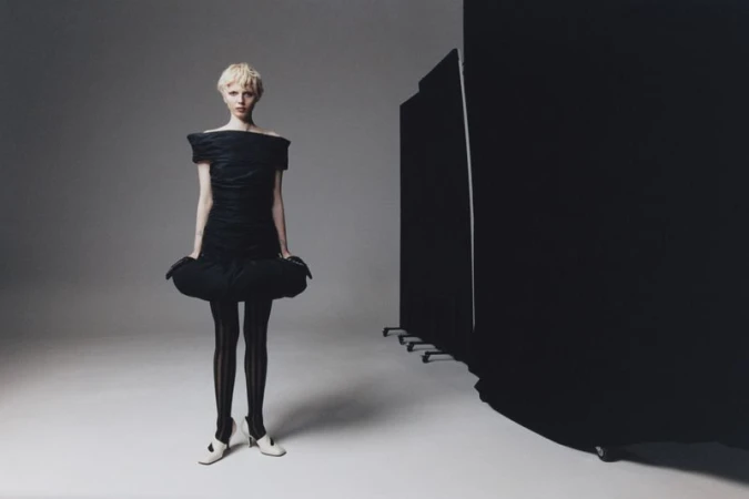 Fashion shot of a model in a black dress and striped tights, posing in a photo studio with a dark screen backdrop. 