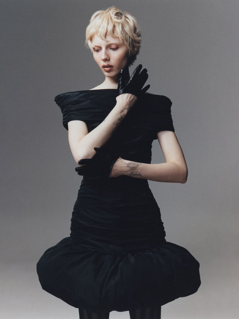 Elegant woman with short blond hair in a black off-the-shoulder dress and gloves posing in a studio, fashion photography.