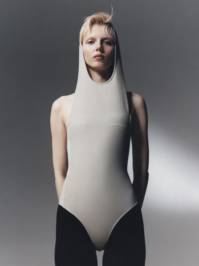 Woman with short blonde hair poses in a studio wearing a beige bodysuit with a hood and black tights against a grey background.