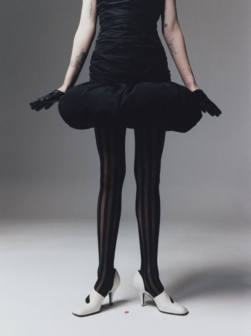 Fashionable woman in a black dress and striped tights stands in a photo studio, wearing white heels and black gloves against a gray backdrop.