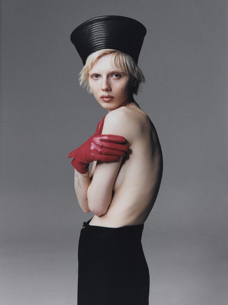Portrait of a slender fair-skinned adult woman with blonde hair, black skirt, red gloves, and black headwear against a grey studio backdrop.