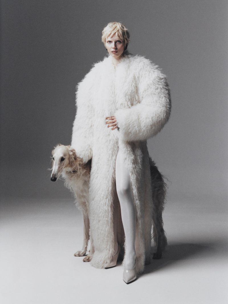A fair-skinned woman with blonde hair poses in a shaggy white coat next to a Borzoi dog against a neutral grey backdrop in studio lighting.