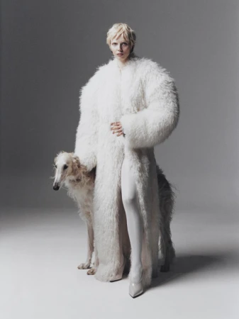 A fair-skinned woman with blonde hair poses in a shaggy white coat next to a Borzoi dog against a neutral grey backdrop in studio lighting.