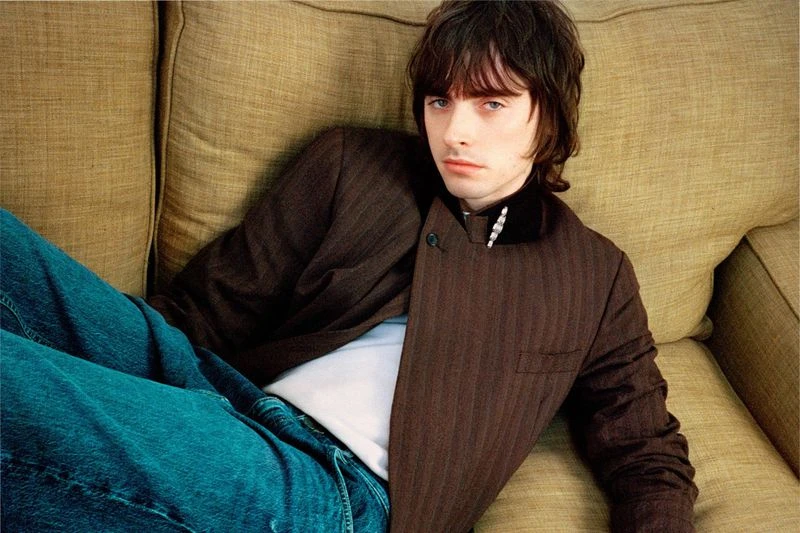 Dark-haired man reclines on a tan sofa wearing a pinstripe jacket, t-shirt, and jeans in a portrait-style studio image.