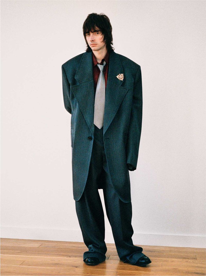 A man in an oversized teal suit stands against a white wall, creating a striking fashion portrait with a vintage aesthetic.