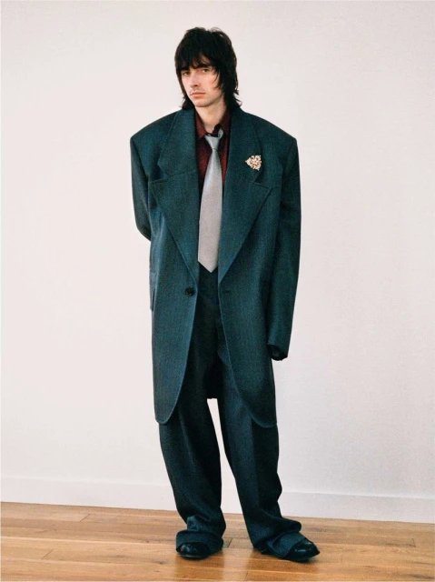 A man in an oversized teal suit stands against a white wall, creating a striking fashion portrait with a vintage aesthetic.