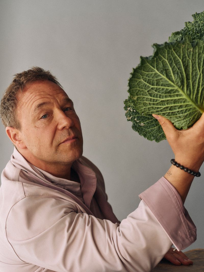Portrait of a fair-skinned adult male holding a large green cabbage against a neutral background, wearing a pale pink shirt.
