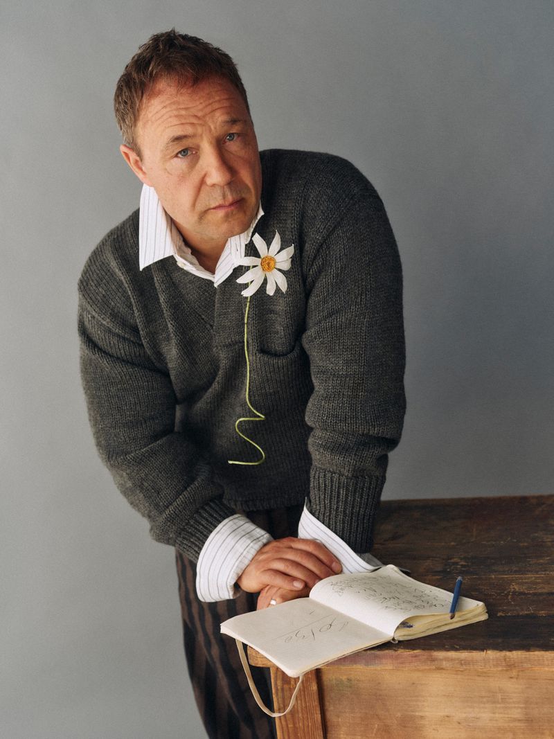 Adult man poses indoors with gray sweater, collared shirt, and brown pants, resting arms on desk next to open notebook and pen.