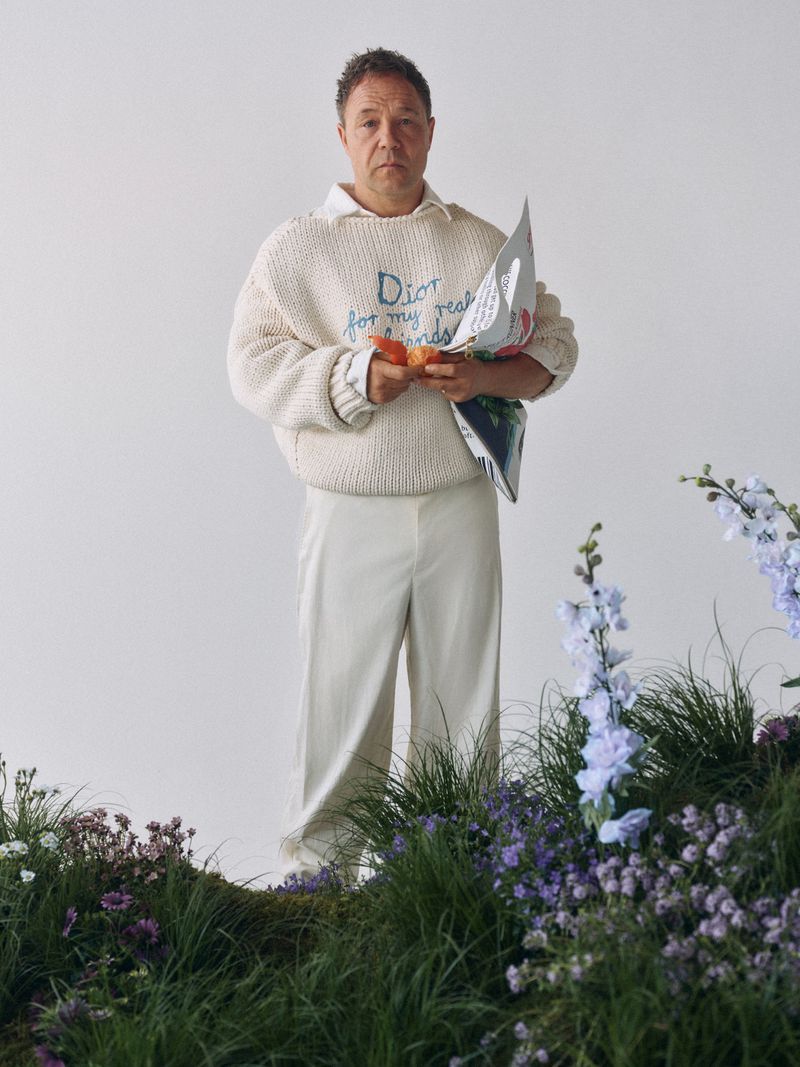 Adult male stands in a cream sweater and pants holding a carrot and newspaper, surrounded by flowers in a photo studio setting.