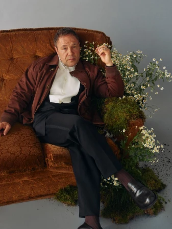 Portrait of an adult male sitting in a brown chair in a studio setting with greenery and a neutral backdrop, wearing a brown jacket.