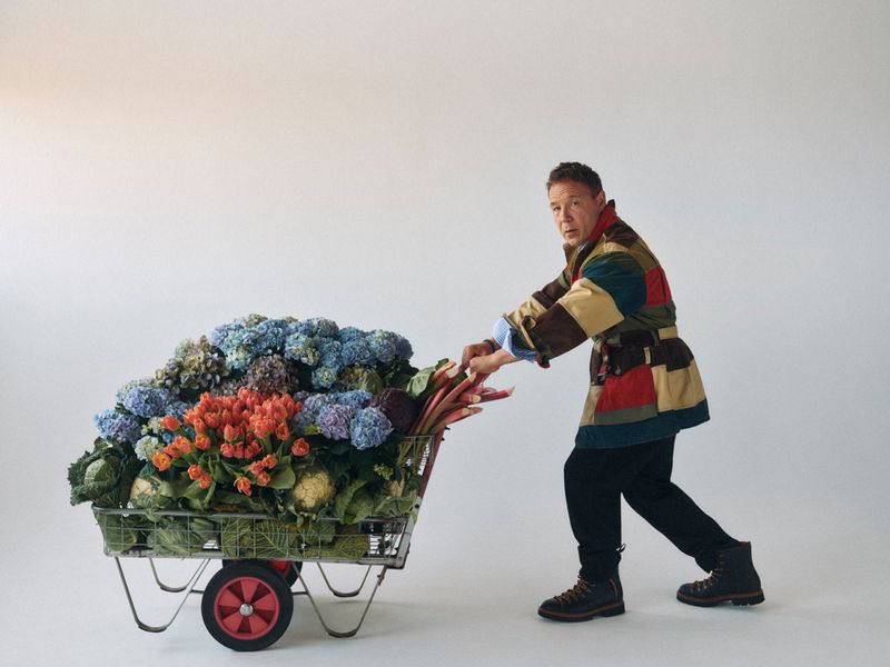 Adult male pushing a shopping cart filled with flowers and vegetables in a light studio against a white seamless backdrop.