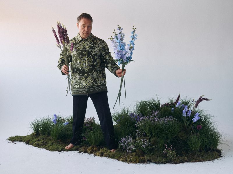 Man stands barefoot on mossy ground holding purple and blue flowers in a studio setting with a white background. He is wearing a patterned shirt.