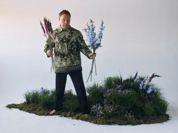 Man stands barefoot on mossy ground holding purple and blue flowers in a studio setting with a white background. He is wearing a patterned shirt.