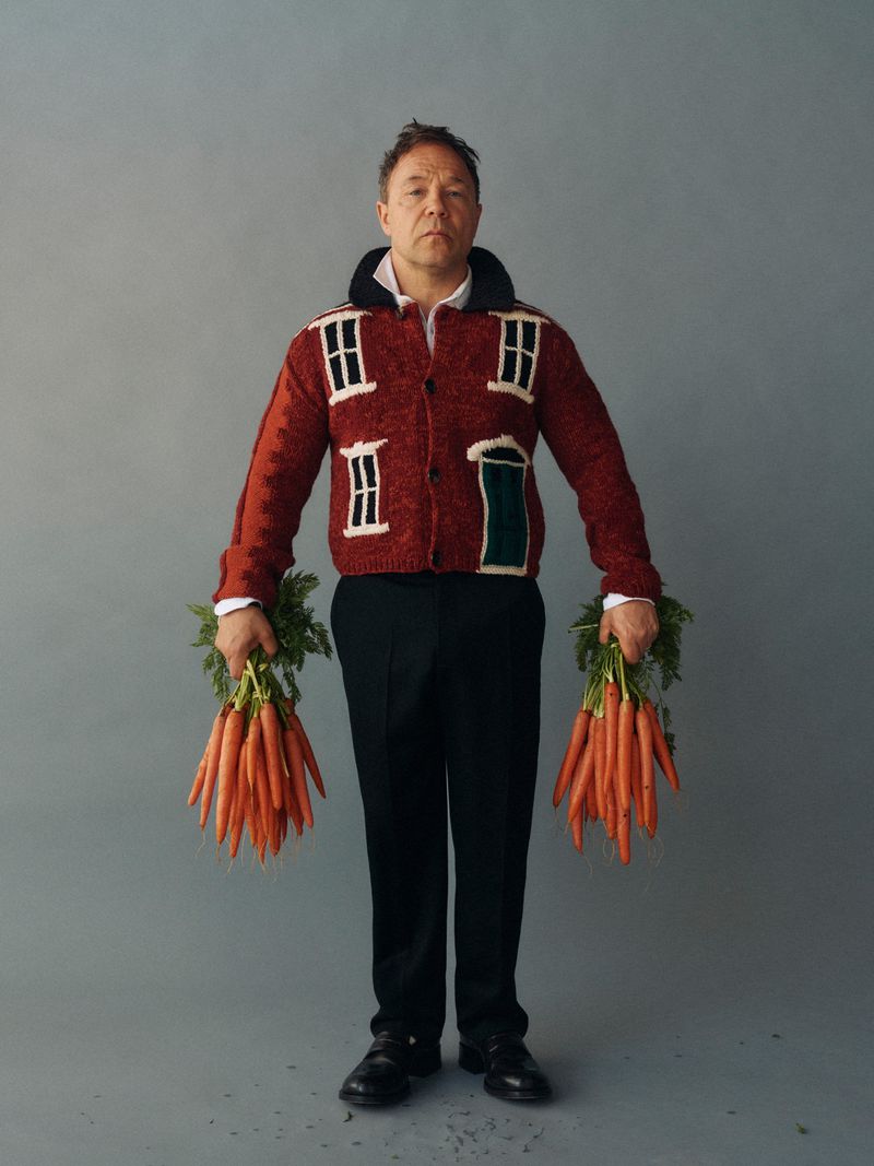 Man in house-themed sweater holds carrots in studio, black pants and dress shoes, set against a grey backdrop. Food and fashion crossover.
