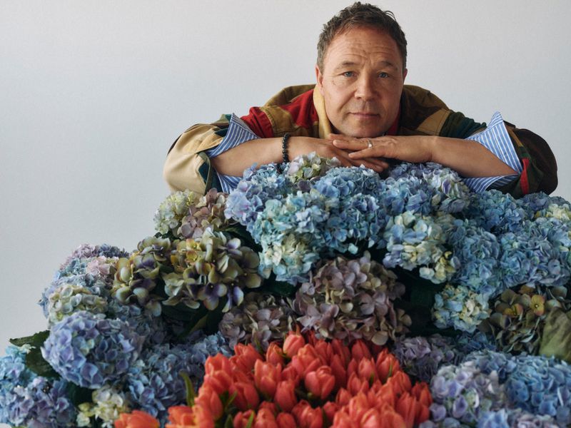 Portrait of a mature adult male leaning on a large arrangement of blue hydrangeas and orange tulips, indoors with a white background.