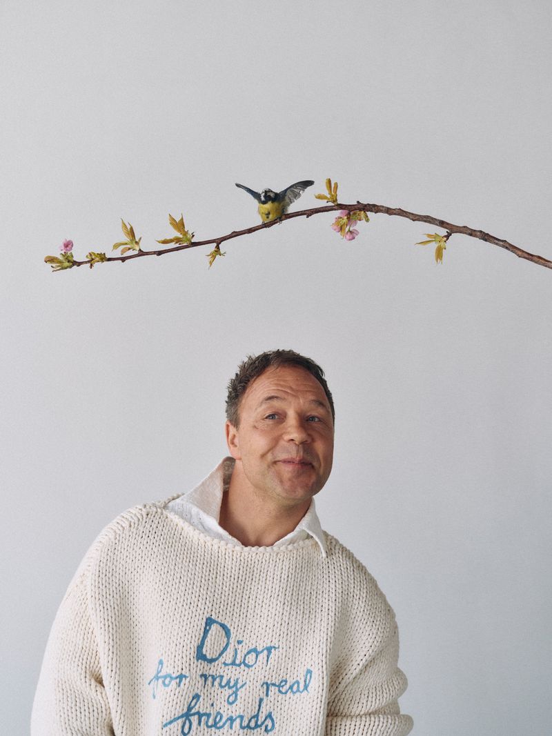 Portrait of a smiling man in a sweater with a bird perched on a branch above him, set against a neutral studio background.