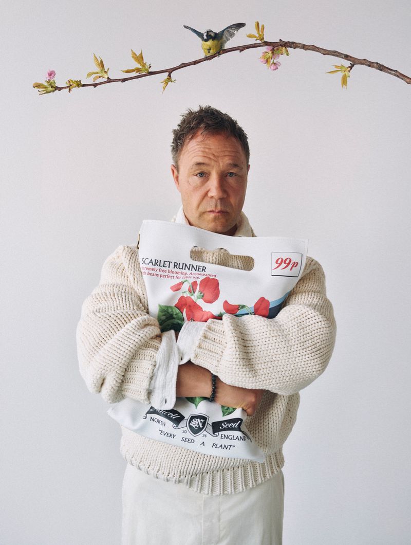 Adult male in a cream sweater and white skirt holds a bag of scarlet runner seeds, set against a plain white background with tree branch.