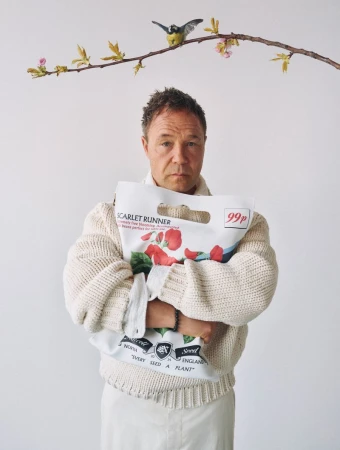 Adult male in a cream sweater and white skirt holds a bag of scarlet runner seeds, set against a plain white background with tree branch.