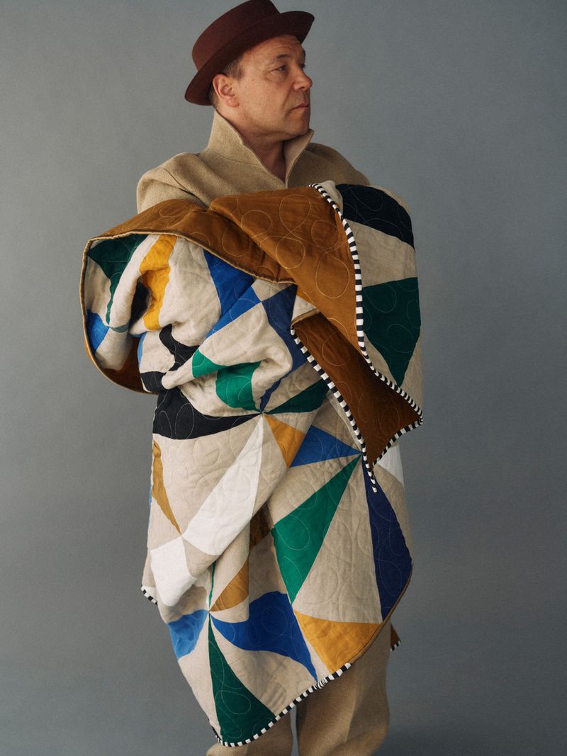 Adult man wrapped in a geometrically patterned quilt in a studio setting, wearing a hat and overcoat against a grey background.
