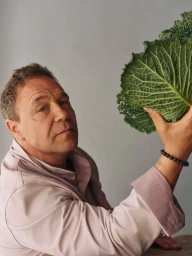 Close-up portrait of middle-aged man in light pink shirt holding a large green leafy cabbage against a plain grey studio backdrop.