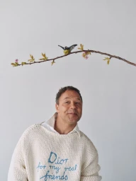 Man wearing a cream knit sweater with blue embroidered text beneath a flowering twig with a small bird against a white background