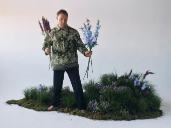 Man standing on a grassy patch holding bundles of purple and blue flowers against a white background for GQ UK.