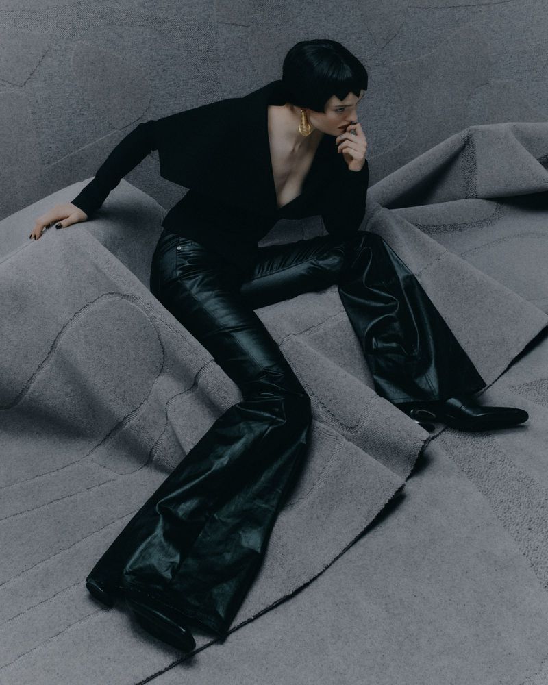 A woman poses in a black outfit on a gray textured surface. She wears shiny black pants, a black top, and gold earrings.