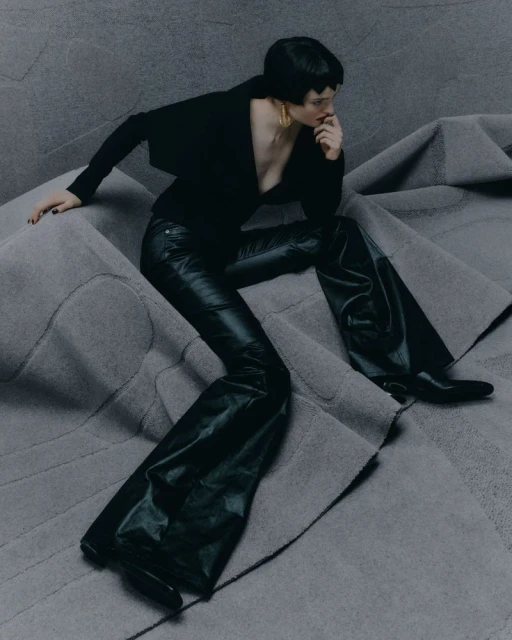 A woman poses in a black outfit on a gray textured surface. She wears shiny black pants, a black top, and gold earrings.