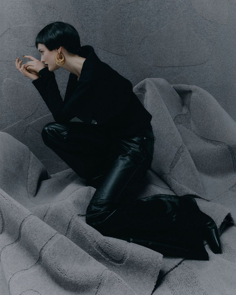 A woman with short dark hair poses in black clothes, including a top, leather pants, and boots, against a textured grey background for a fashion shoot.