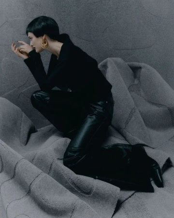 A woman with short dark hair poses in black clothes, including a top, leather pants, and boots, against a textured grey background for a fashion shoot.