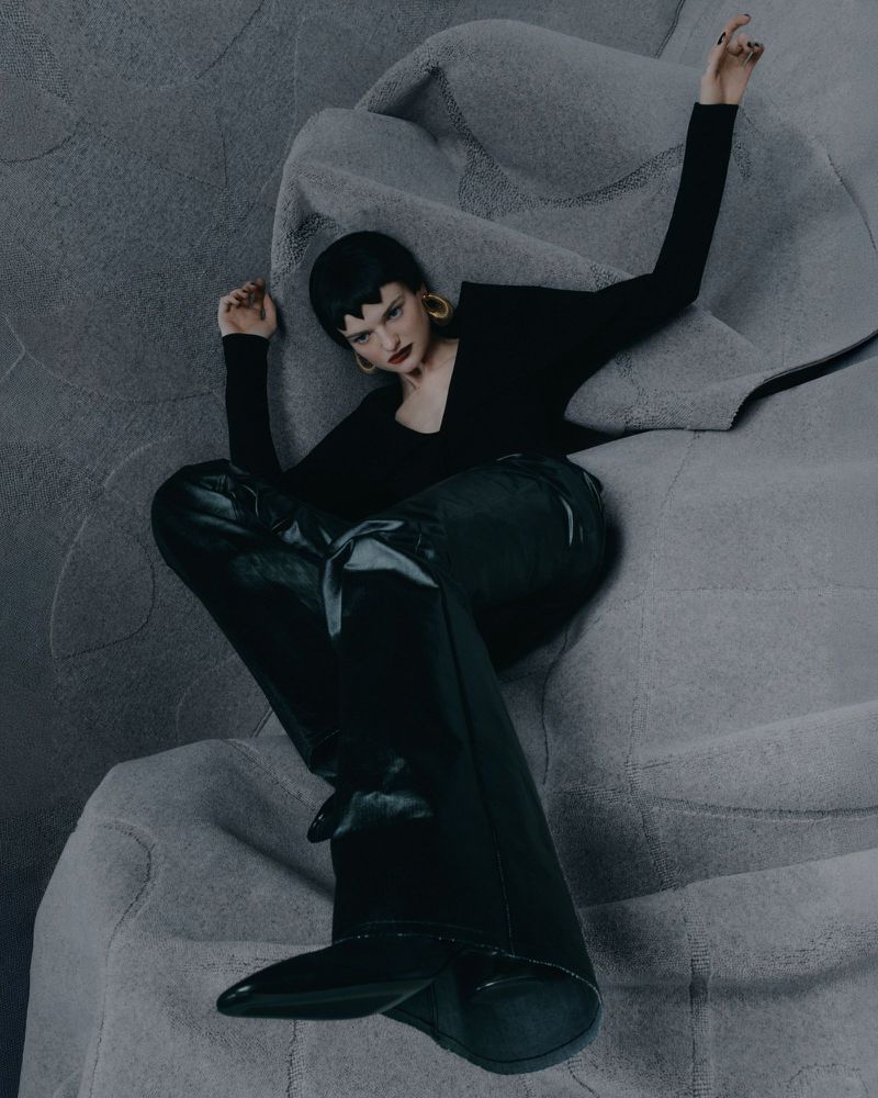 Fashion shot of a woman with short dark hair in black top, pants, and boots sitting on a gray fabric surface, looking cool and collected.
